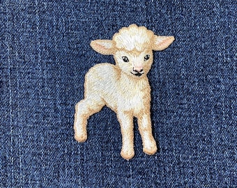 May include: Embroidered patch featuring a cream-colored lamb with a fluffy head, standing on four legs. The lamb has black eyes and a black nose. The patch is set against a dark blue denim background.