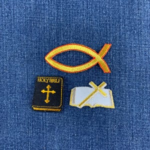 Peut inclure: Trois écussons brodés représentant des symboles religieux : un contour de poisson jaune, un livre noir avec le texte HOLY BIBLE et une croix jaune, et un livre blanc ouvert avec une croix jaune, disposés sur un tissu en jean bleu.