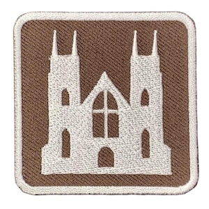Könnte beinhalten: Ein quadratischer gestickter Aufnäher mit der weißen Silhouette einer Kirche mit zwei hohen Türmen und einem zentralen Bogenfenster mit Kreuz, vor einem einfarbigen braunen Hintergrund mit einem weißen gestickten Rand um den gesamten Umfang.