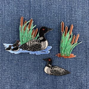 May include: Three embroidered patches featuring common loons. One patch shows an adult loon with two chicks swimming near green cattails on blue water. A second patch shows a single adult loon. A third patch shows a cluster of green cattails.