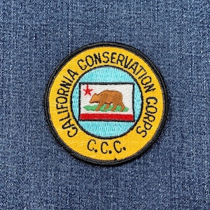 May include: A circular patch with a yellow border and black trim. The text "CALIFORNIA CONSERVATION CORPS" is in a yellow arc around the top and right side. The center features the California state flag with a bear and the letters "C.C.C."