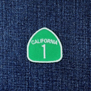 May include: A green embroidered patch shaped like a shield with a white border. The patch features the white text CALIFORNIA arched over the large white number 1. The patch is displayed against a dark blue denim fabric background with visible texture.