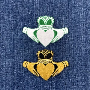 May include: Two embroidered Claddagh patches featuring a heart held by two hands topped with a crown. One patch is white with green accents and a green crown, while the other is gold with black accents and a black crown, both stitched on denim.