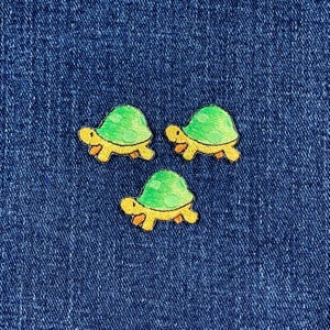 Könnte beinhalten: Drei gestickte Schildkröten-Patches mit grünen Panzerungen und gelben Körpern auf blauem Denim-Hintergrund. Die Schildkröten haben orangefarbene Füße und blicken in verschiedene Richtungen. Geeignet für Kleidung oder Bastelprojekte.