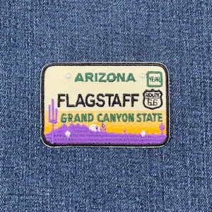 Könnte beinhalten: Ein weißer und gelber gestickter Aufnäher mit dem Text "Arizona Flagstaff Grand Canyon State" und einem Route 66 Schild. Der Aufnäher hat eine violette Bergkette und grüne Kakteen.