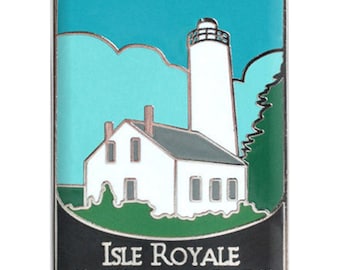 May include: A rectangular enamel pin featuring a white lighthouse and building against a teal sky and green landscape. The pin has the text "Isle Royale NATIONAL PARK" in white letters on a black background.