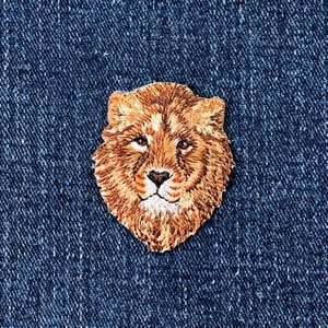 May include: Embroidered patch featuring a detailed lion's head in shades of brown and tan. The patch is sewn onto a blue denim fabric, showcasing the intricate threadwork and design. The lion's face is centered, with a heart-shaped nose.