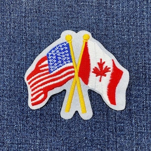 May include: Embroidered patch featuring the flags of the United States and Canada crossed. The American flag has red and white stripes with a blue field of white stars. The Canadian flag is red with a white maple leaf.