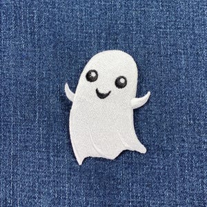 May include: A white embroidered patch in the shape of a ghost with two small arms extended outward. The ghost features two round black eyes and a small black curved mouth, creating a simple facial expression on the white fabric surface.