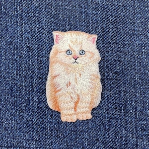 以下が含まれることがあります： 青い目をしたふわふわの薄いオレンジ色の猫の刺繍パッチ。猫は直立して座っています。パッチは、濃い青色のデニムの背景に対して、さまざまなオレンジとピンクの色合いで詳細に描かれています。猫の表情は穏やかで好奇心旺盛です。