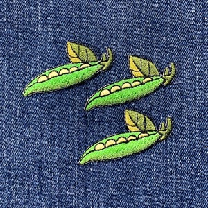 May include: Three embroidered pea pod patches featuring bright green pods with visible yellow peas inside and a small green leaf attached to the top stem. The patches are arranged in a diagonal pattern against a dark blue denim fabric background.