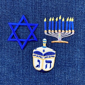 May include: Embroidered patches depicting Jewish symbols: a blue Star of David, a menorah with lit candles, and a dreidel with Hebrew letters. The patches are in blue, white, and yellow, on a denim background.