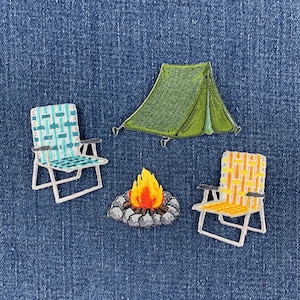 May include: A set of four embroidered patches featuring a green tent, a campfire with orange and yellow flames surrounded by gray stones, a light blue woven folding chair, and a yellow woven folding chair, all arranged on a blue denim fabric.