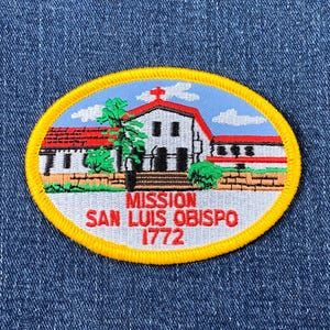 May include: Yellow embroidered patch with a white building with red roof and a blue sky. The text reads "Mission San Luis Obispo 1772".