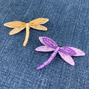 May include: Two embroidered dragonfly patches, one in a light golden yellow and the other in a vibrant purple. Each dragonfly features intricate wing detailing with a sheer, lace-like texture and a slender, segmented body design.