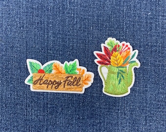 May include: Two embroidered patches featuring autumn themes. One patch is a rectangular wooden sign with the text Happy Fall surrounded by green and orange leaves. The second patch is a green watering can filled with red, yellow, and orange leaves.