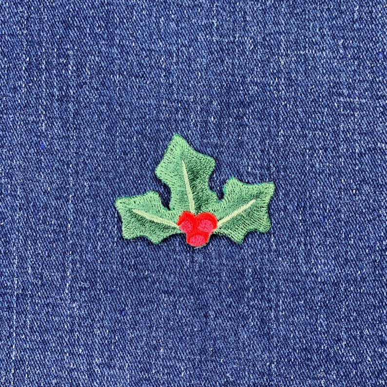 May include: A green embroidered holly leaf with red berries, set against a blue denim background. The holly design is detailed with light green stitching, creating a festive and seasonal embellishment.