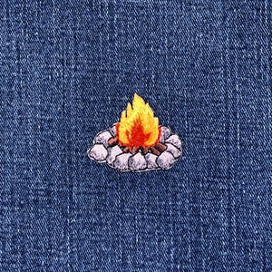 May include: Embroidered patch featuring a campfire design. The patch shows a campfire with orange and yellow flames, surrounded by gray stones and brown logs. The patch is on a blue denim background.
