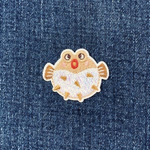 May include: Embroidered patch with a cartoon pufferfish design. The patch has a tan head, white body with gold spikes, and tan fins. The pufferfish has large, round eyes and a red mouth. The patch is on a blue denim background.