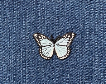 May include: A light blue butterfly patch with white and black detailing, set against a textured denim background. The butterfly has a delicate design, perfect for adding to clothing or accessories. The patch is approximately 5 cm wide.