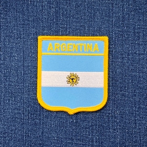 May include: Embroidered patch featuring the Argentinian flag design. The patch has a light blue background with a white horizontal stripe and a yellow sun emblem. The word "ARGENTINA" is embroidered in yellow at the top, with a yellow border.