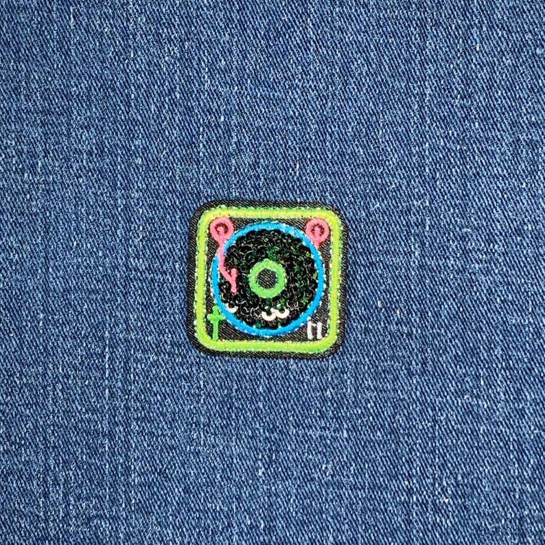 May include: A square patch featuring a colorful turntable design. The patch has a black center with a green circle, surrounded by a blue circle and pink accents. The patch is outlined in green and has small white details.
