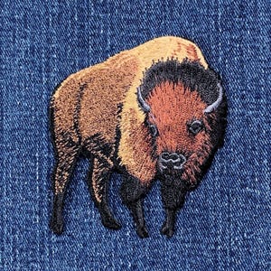 May include: Embroidered patch of a brown and black bison with a dark brown mane. The bison is standing on a blue denim background.