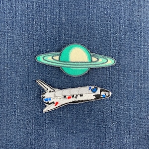 May include: Two embroidered patches featuring space themes. The top patch is a green and cream planet with rings. The bottom patch is a white, black, and blue space shuttle with small red accents. Both patches are detailed with visible stitching.