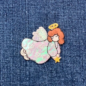 May include: A pink and iridescent angel with a halo and a star in its hand. The angel is embroidered on a blue denim fabric.
