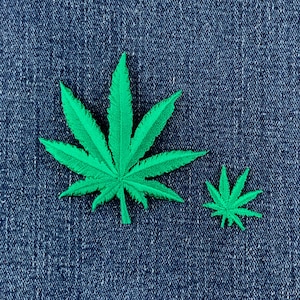 May include: Two embroidered patches in the shape of seven-pointed cannabis leaves. The patches are a vibrant, solid green color with visible thread texture and serrated edges, displayed against a dark blue denim fabric background.