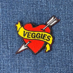 May include: A red heart with a yellow banner that says "VEGGIES" is pierced by a white arrow. The heart and banner are embroidered on a blue denim background.
