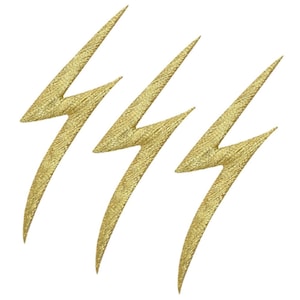 May include: Three gold-coloured lightning bolt-shaped embellishments with a textured surface. The bolts are arranged diagonally against a white background. These decorative elements could be used for crafts or fashion.