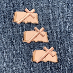 May include: Three embroidered patches featuring a cross laid over an open book. The patches are in shades of pink and tan, with a detailed stitched design. The patches are arranged on a blue denim background.