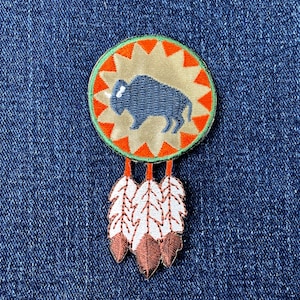 May include: Embroidered patch with a blue bison design within a tan circle with orange starbursts, a green border, and three hanging feathers. The feathers are white with brown tips. The patch is on a blue denim background.