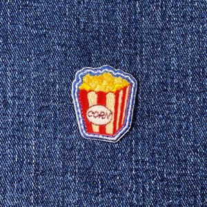 May include: A colorful popcorn patch with a red and white striped container, yellow popcorn, and the word "CORN" in a white oval. The patch has a blue border and is set against a blue denim background.