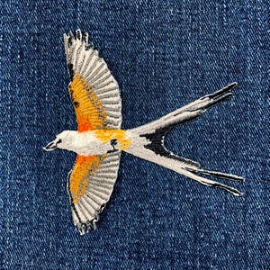 以下が含まれることがあります： 白、オレンジ、黒のディテールが施された、飛行中の鳥の刺繍パッチ。鳥は翼を広げ、長い二股の尾を持っています。パッチは青いデニムの背景に配置されています。