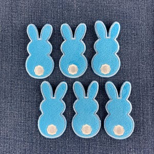 May include: Six bright blue felt bunny rabbit appliques arranged in two rows. Each bunny shape features long ears, a rounded body, and a small, circular white tail detail at the base. The edges of each bunny are finished with a light blue stitched border.