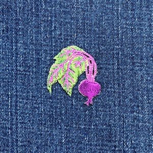 May include: Embroidered patch featuring a stylized beet design. The beet has a vibrant pink root and stem, with green and pink leaves. The patch is set against a textured blue denim background.