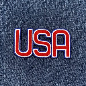 May include: Embroidered patch with the letters "USA" in red with a white border and blue outline, set against a blue denim background. The patch is a patriotic emblem.