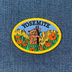 May include: A yellow and blue embroidered patch with the word "Yosemite" in black lettering. The patch features a brown bear standing on its hind legs, holding a tree trunk. The background includes orange flowers and green foliage.