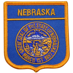 May include: A blue and gold embroidered patch featuring the word "NEBRASKA" at the top. The center displays the state seal with text "SEAL OF THE STATE OF NEBRASKA" and "MARCH 1ST 1867". The patch has a gold border.