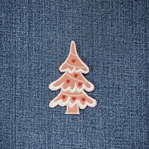 May include: A light pink Christmas tree patch with white trim and small red heart accents. The patch is shaped like a stylized tree and is set against a blue denim background.