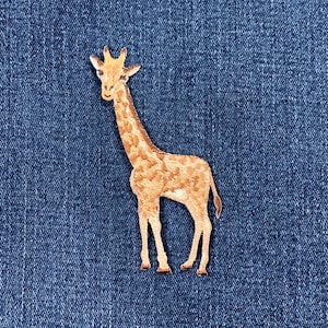 May include: A giraffe-shaped patch with a detailed embroidered design in shades of brown and tan. The patch features a long neck, small horns, and a spotted pattern. The patch is likely for clothing or accessories.