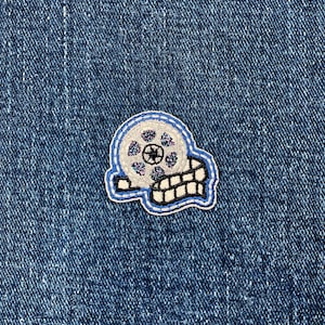 May include: A film reel patch with a blue outline and a white and gray center. The patch is on a blue denim background. The film reel has a black center and a white and gray film strip.