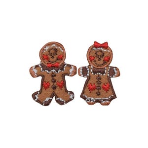 May include: Two embroidered patches shaped like gingerbread figures. One figure wears a red bow tie, and the other wears a red hair bow. Both feature brown fabric, white icing-style stitching, and red heart accents on their torsos and cheeks.