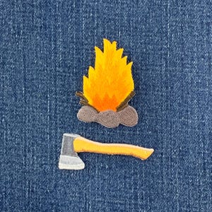 May include: An embroidered patch featuring a bright yellow and orange campfire with gray stones at the base, positioned above a separate embroidered patch of a gray axe head with a light orange handle, both displayed on a blue denim fabric.