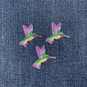 May include: Three embroidered hummingbird patches featuring purple wings, green bodies, and tan underbellies. Each bird is depicted in a flying position with wings extended upward, showcasing detailed stitching and vibrant thread colors throughout.