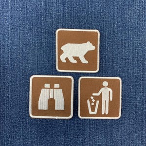 May include: Three square embroidered patches with a brown background and white borders. The top patch features a white bear silhouette. The bottom left patch shows white binoculars, and the bottom right patch depicts a person placing trash in a bin.