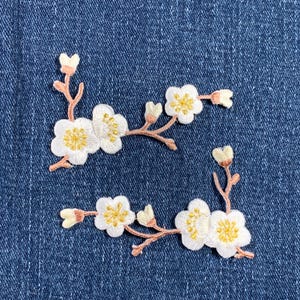 May include: Two embroidered white flower patches with pink stems and yellow centers on a blue denim background.