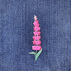 Könnte beinhalten: Ein pinkfarbener, gestickter Blumenaufnäher mit grünen Blättern, vor einem blauen Jeanshintergrund. Der Aufnäher zeigt ein detailliertes Blumendesign, ideal zum Hinzufügen einer dekorativen Note zu Kleidung oder Accessoires.